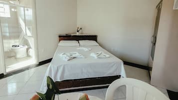 Habitación de hotel con cama, ventilador de techo, baño visible a través de una puerta abierta y una mesa con un jarrón de rosas.