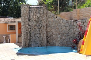 A stone water feature with a cascading waterfall into a blue pool, surrounded by a brick wall and a tiled roof.