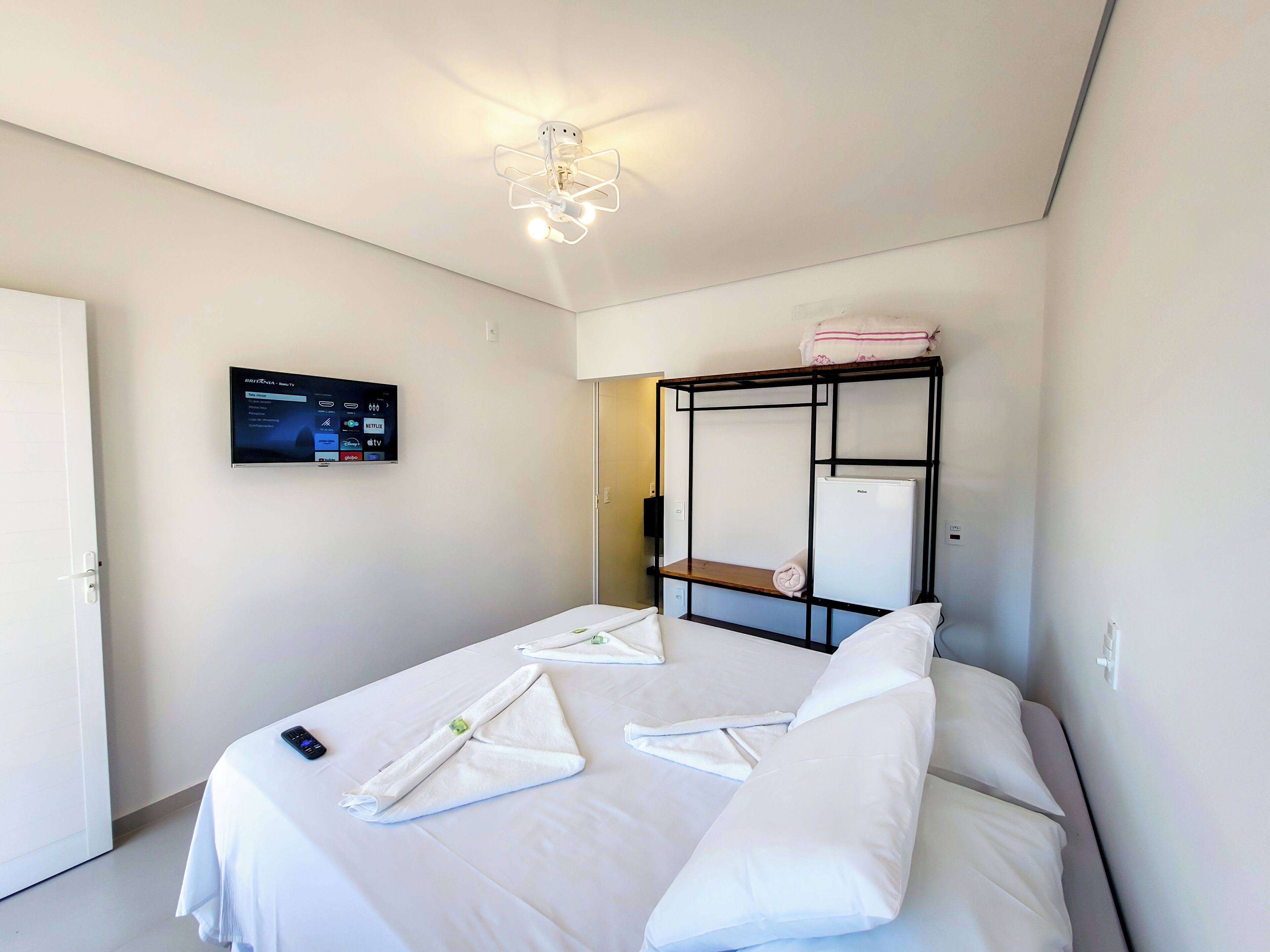 A hotel room with a bed, a flat-screen TV mounted on the wall, a ceiling light fixture, and a small refrigerator.