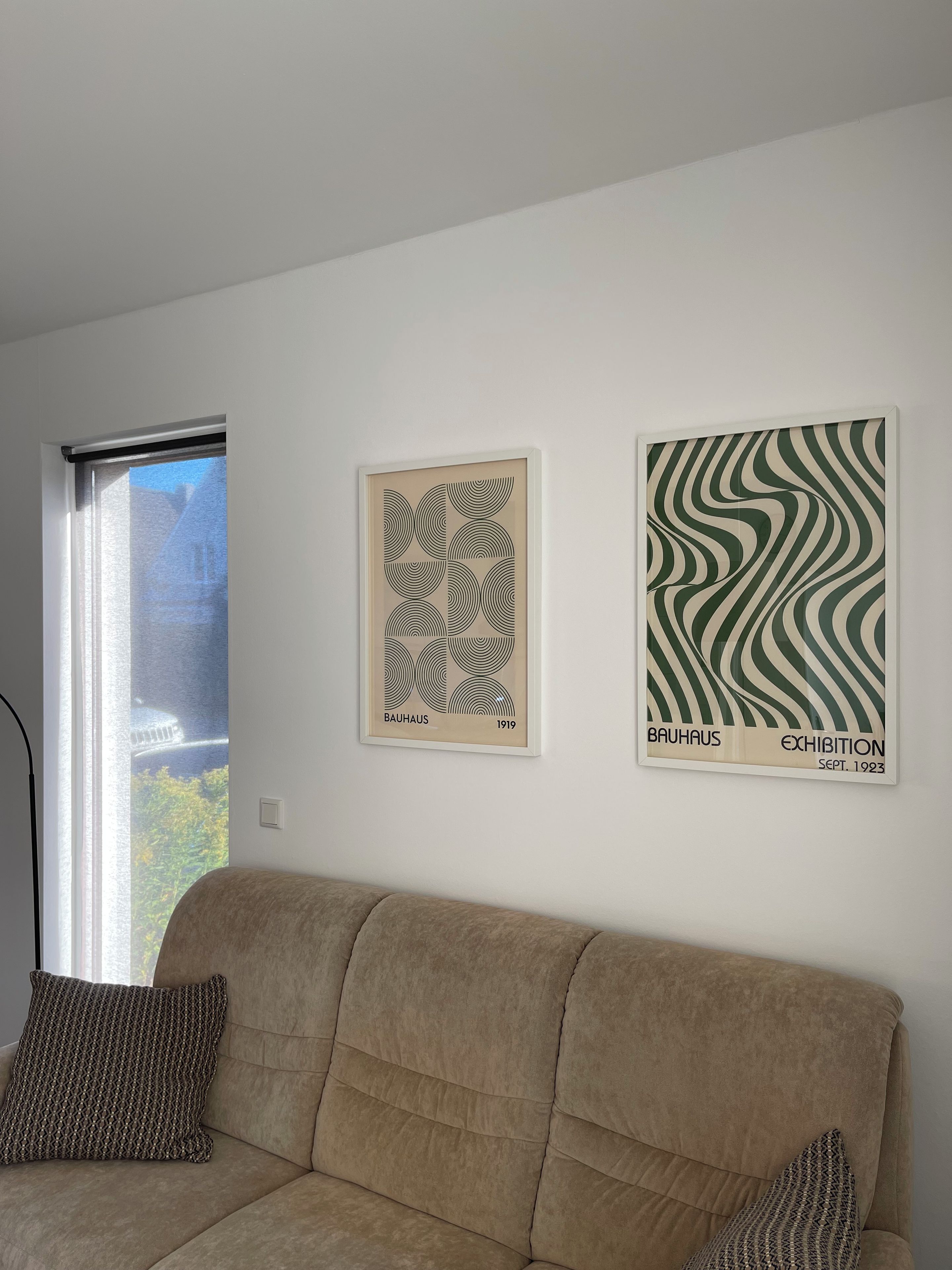 A room with a beige sofa, two framed artworks on the wall, and a window with a view of buildings.