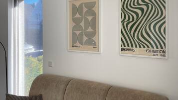 A room with a beige sofa, two framed artworks on the wall, and a window with a view of buildings.
