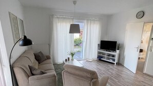 A living room with a sofa, armchair, and a small round table. There is a TV stand with a flat-screen TV and a clock on the wall.