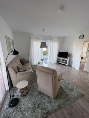 A living room with a sofa, armchair, and a small round table. There is a TV stand with a flat-screen TV and a clock on the wall.