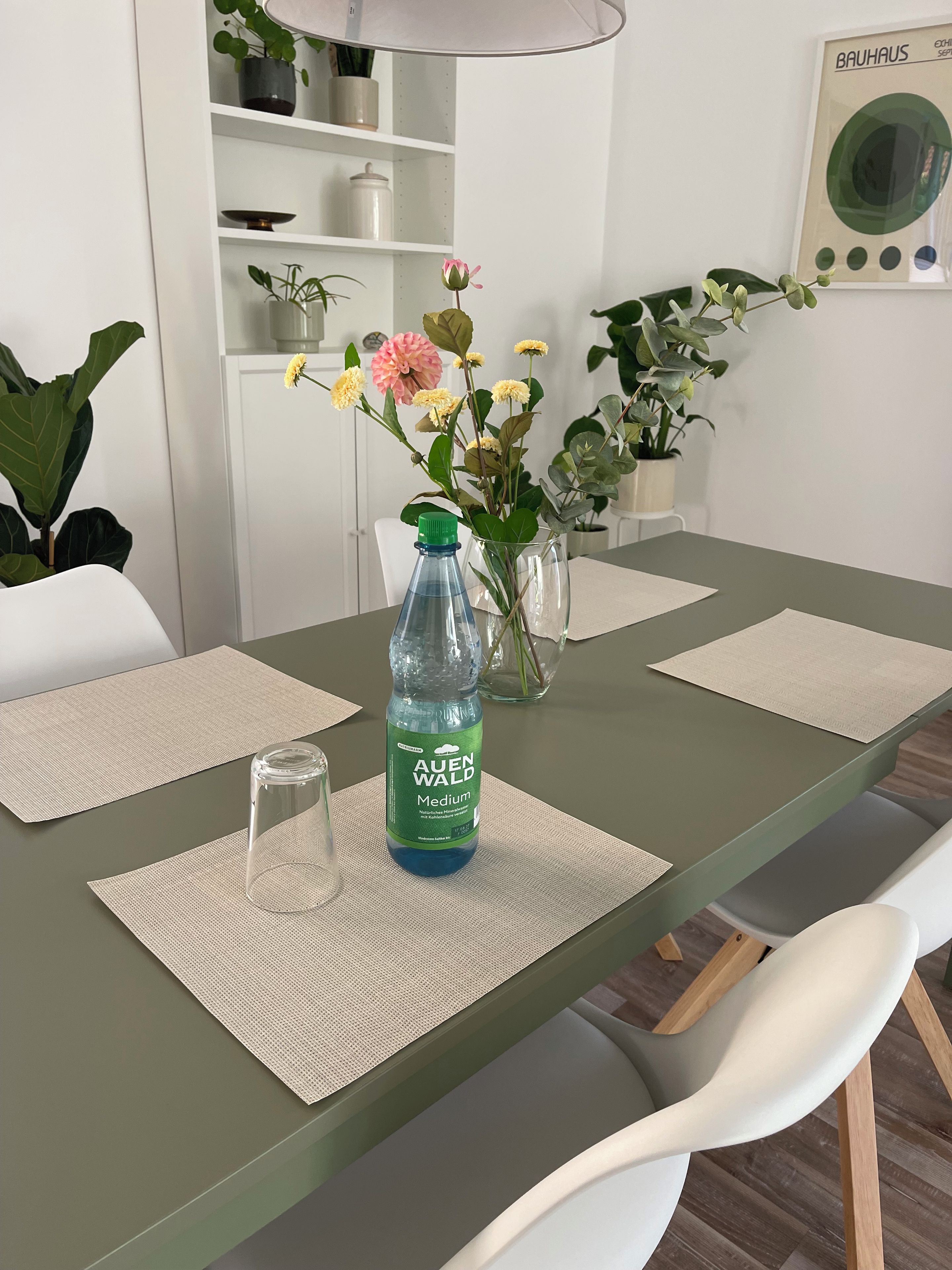 A dining table with a water bottle, a vase with flowers, and a framed picture on the wall.