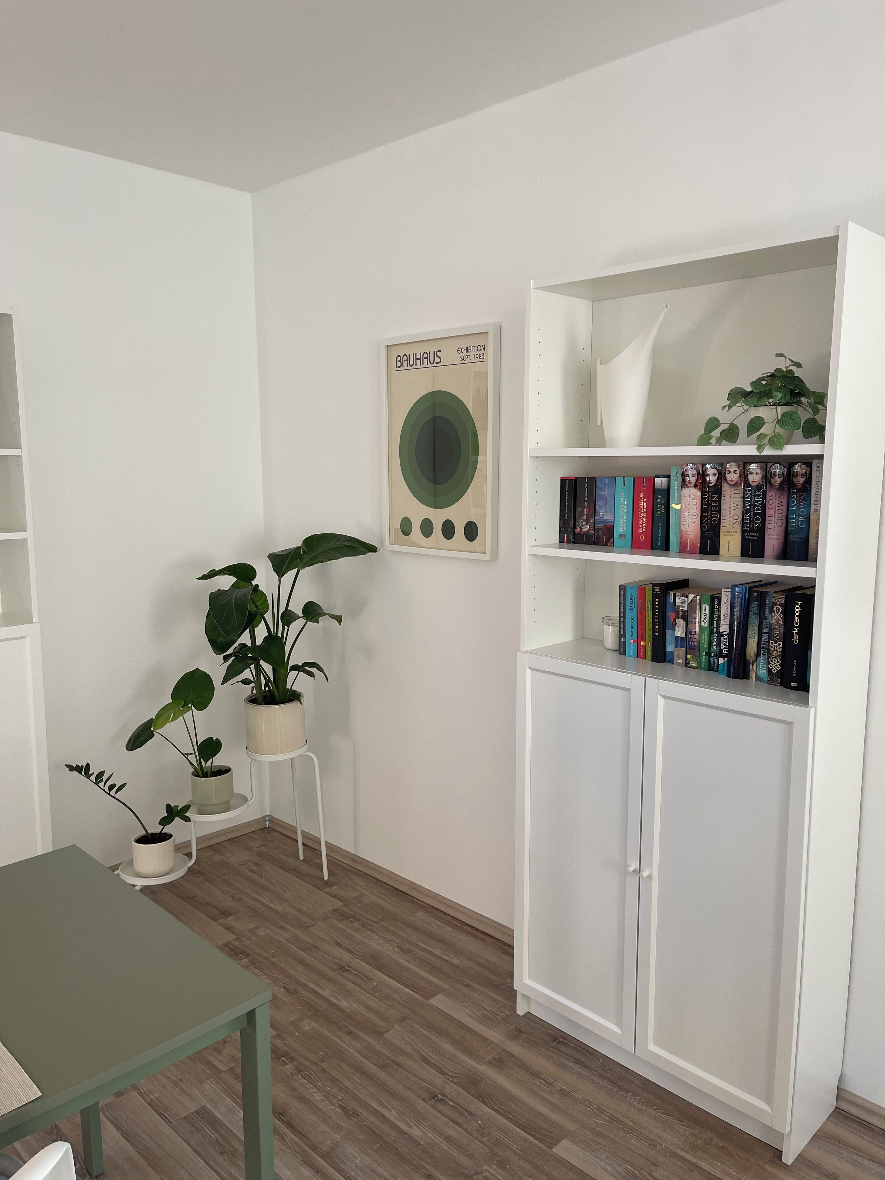Ein Zimmer mit einem weißen Bücherregal, einem Tisch und Topfpflanzen.