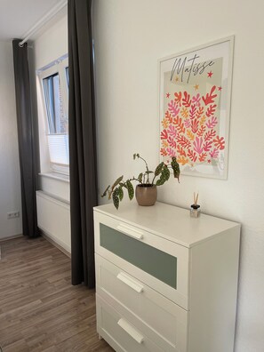 A room with a white dresser, a potted plant, a framed artwork, a window with blinds, and a radiator. - Vacation Rental in Oldenburg Osternburg or for Short-Term Rentals (Oldenburg (Oldb))