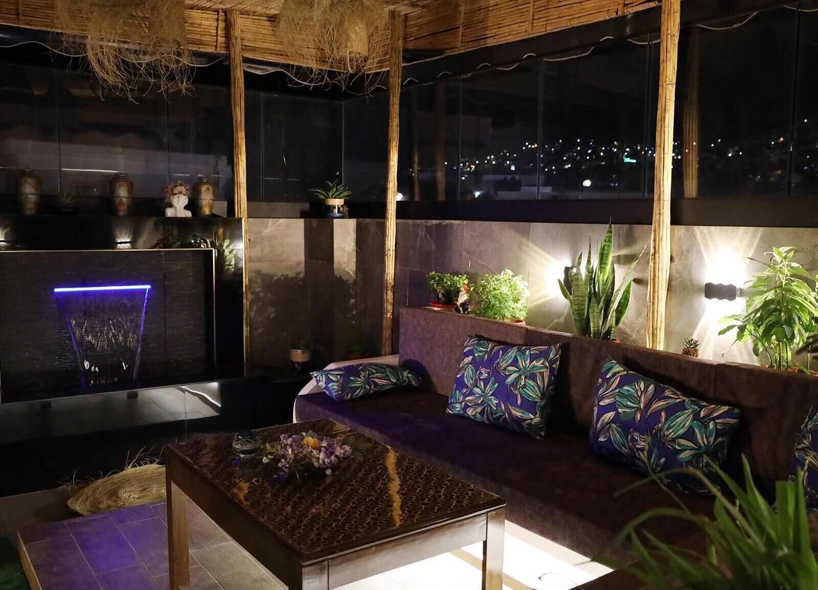 Ein modernes Wohnzimmer mit Sofa, Couchtisch und Kamin mit blauem Licht. An den Wänden stehen Pflanzen und es bietet sich ein Blick auf die beleuchtete Stadt bei Nacht.