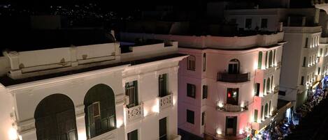 A row of white buildings with balconies, some with lights on, and a street below.