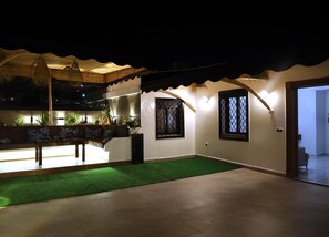 A covered outdoor area with a green artificial turf, a seating area with cushions, and two windows with decorative grills.