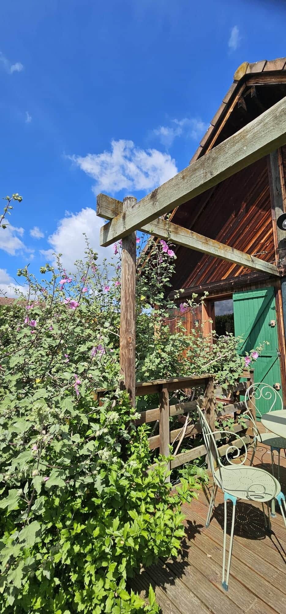 Une terrasse en bois, avec un bâtiment au style rustique en arrière-plan, le tout entouré d’une végétation luxuriante et de plantes en fleurs.