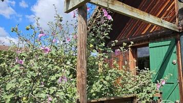 Une terrasse en bois, avec un bâtiment au style rustique en arrière-plan, le tout entouré d’une végétation luxuriante et de plantes en fleurs.