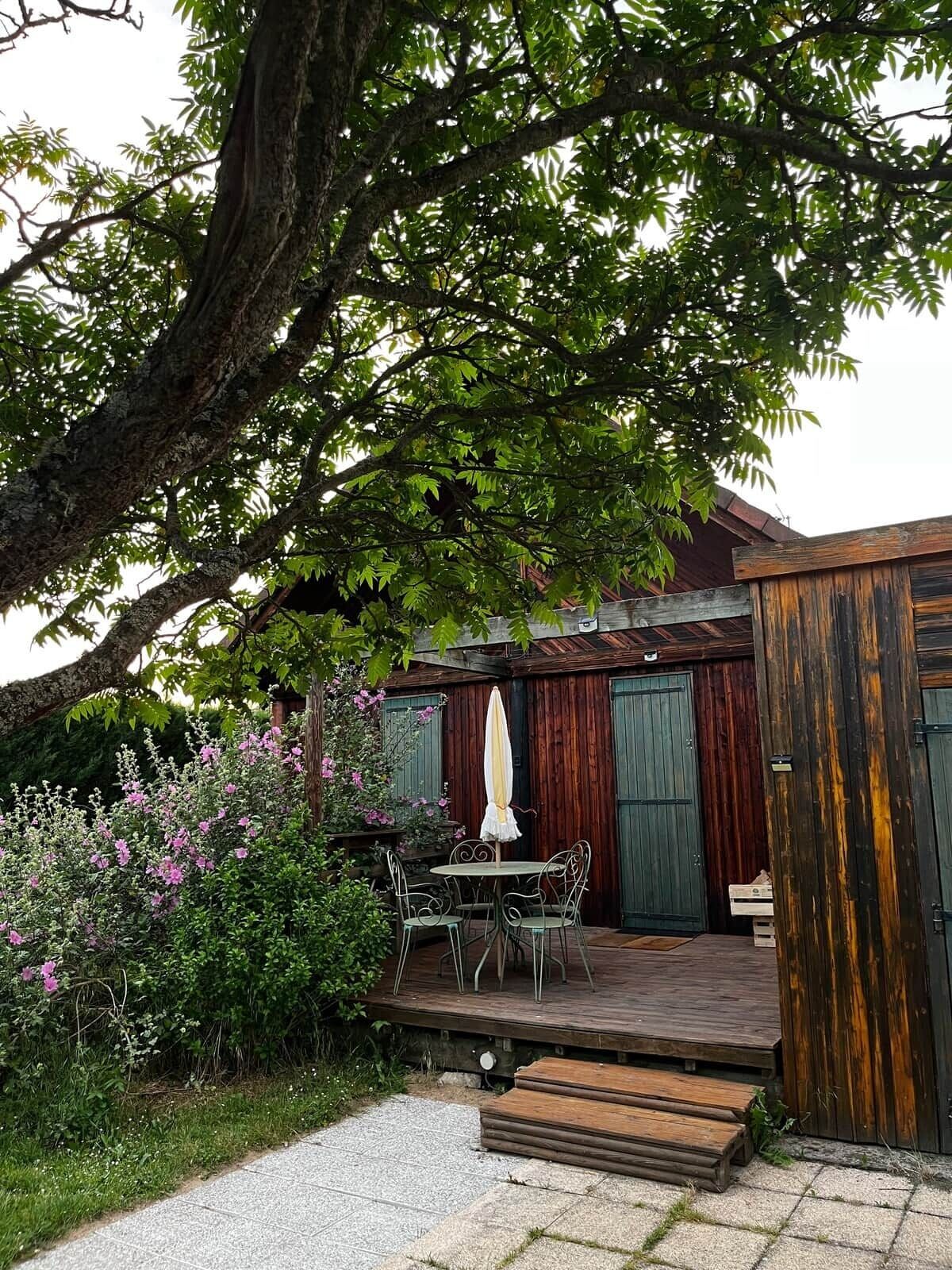 Une terrasse en bois avec une table et des chaises, entourée de verdure et d’un grand arbre.