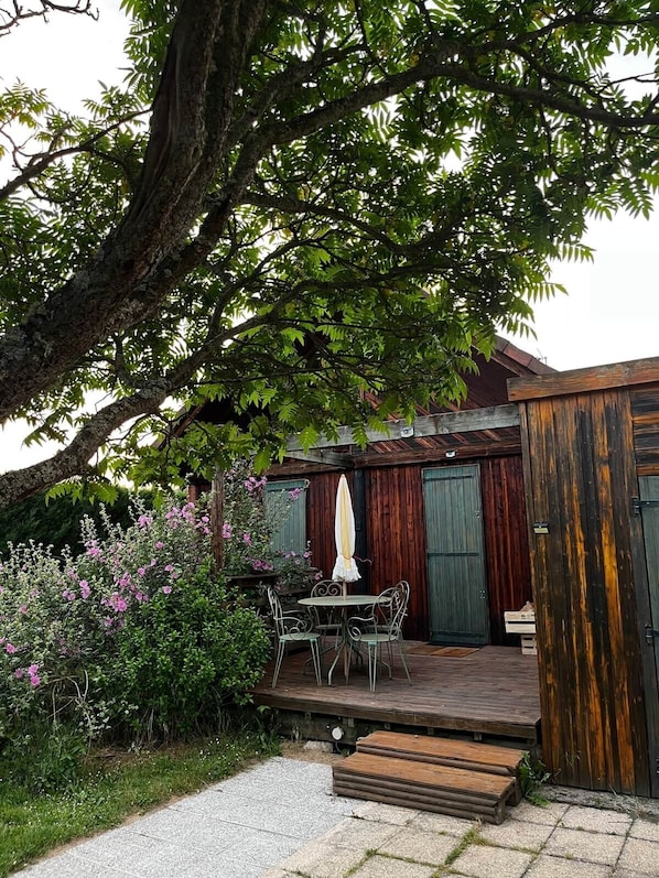 A wooden deck with a table and chairs, surrounded by greenery and a large tree.