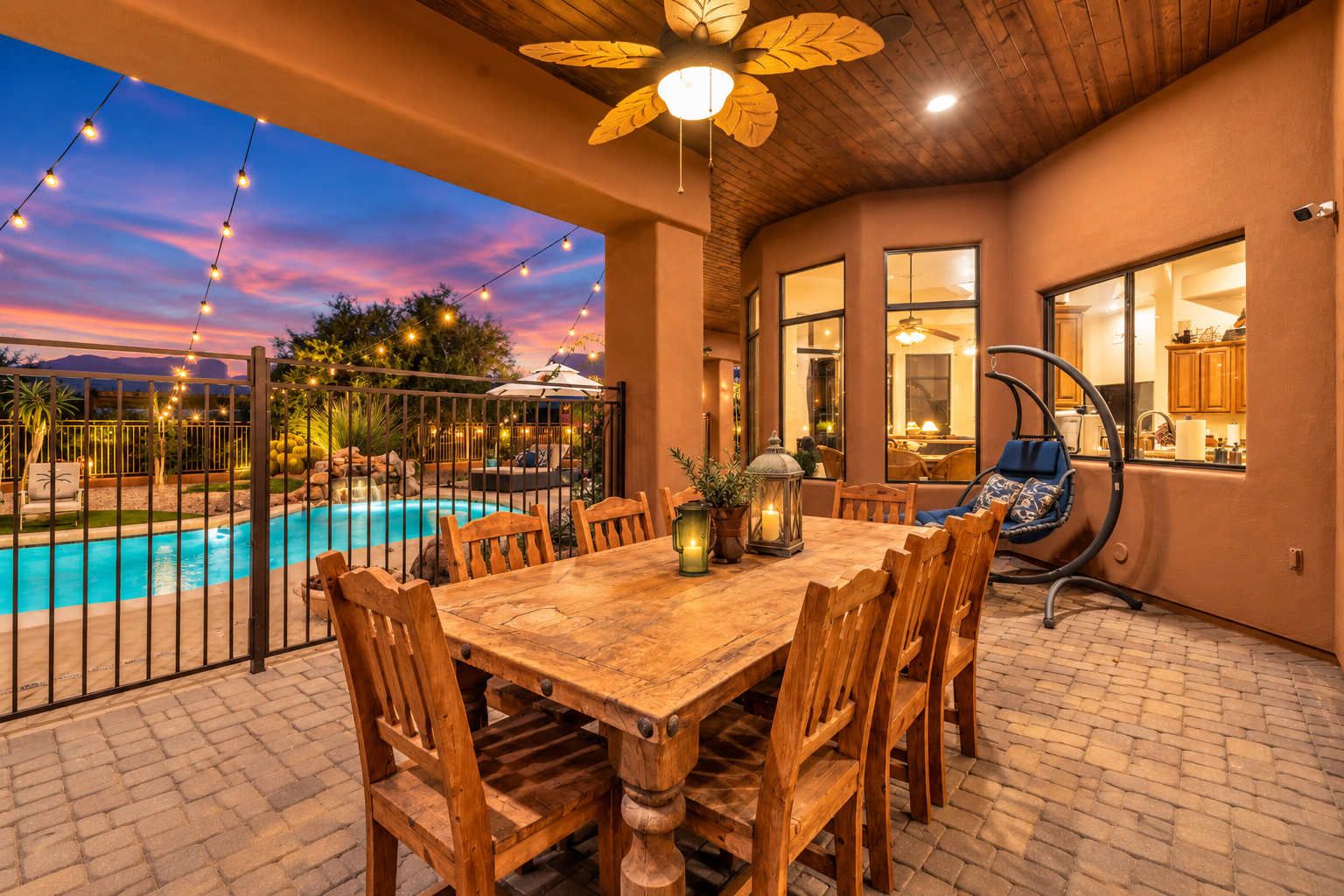An outdoor patio with a wooden table and chairs, a pool, and string lights.