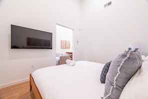 A modern hotel room with a bed, a flat-screen TV mounted on the wall, a small seating area with a sofa and chairs, and a door leading to another room.