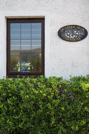 A window with a view of a valley, a sign reading 'QUARTER DECK GARDENS', and a hedge in front.