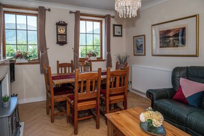 A dining area with wooden chairs and a table, a living room with a sofa, and a view of the outdoors through large windows.