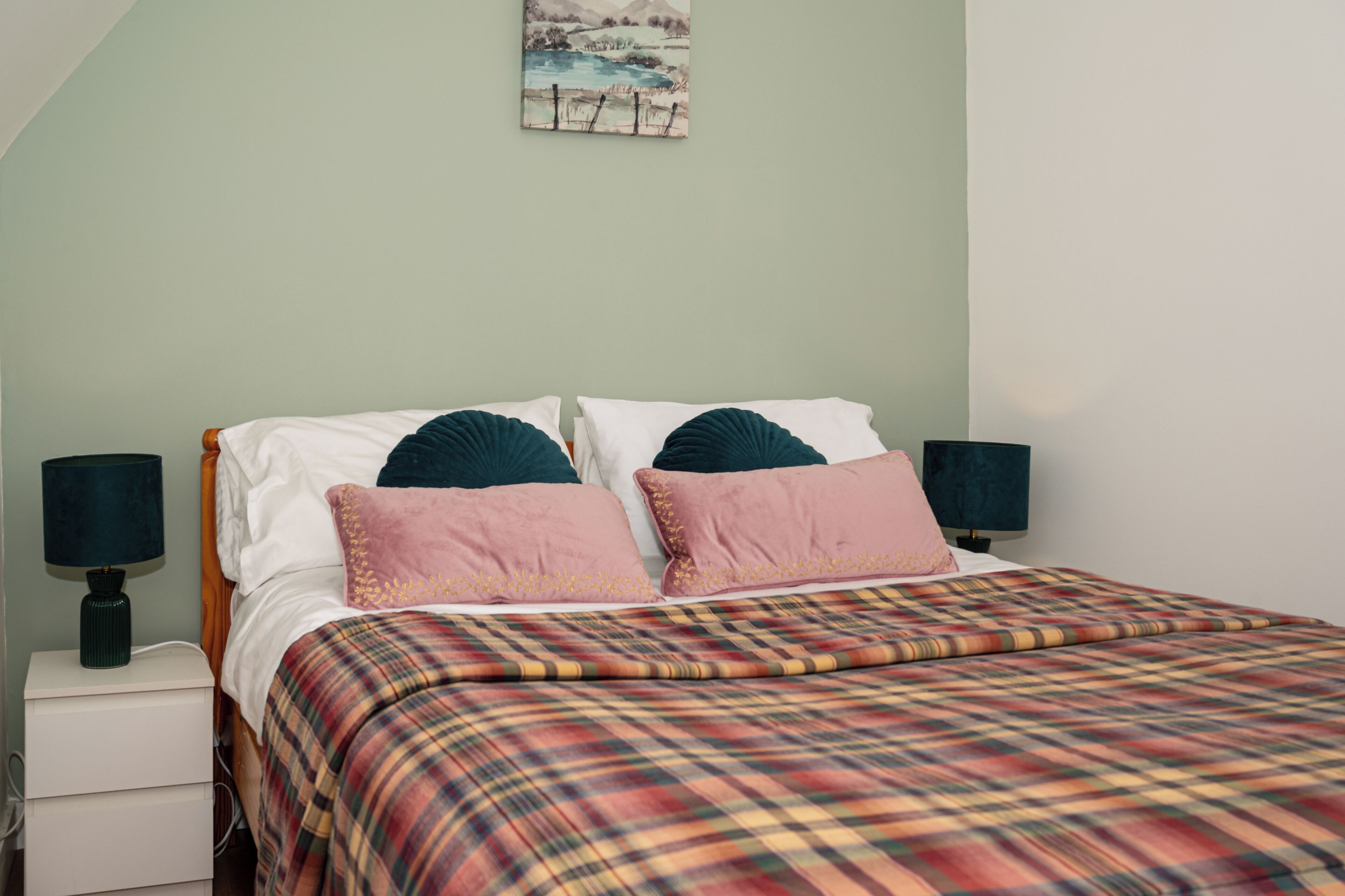 Een bed met een geruite plaid, twee kussens en twee bedlampen.