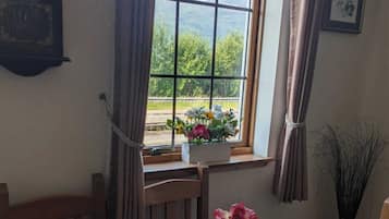 Een eetgedeelte met een houten tafel, stoelen, een vaas met bloemen, een raam met gordijnen en een klok aan de muur.