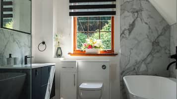 Een moderne badkamer met een vrijstaand bad, een douchecabine met glazen wanden, een toilet en een raam met zonwering.