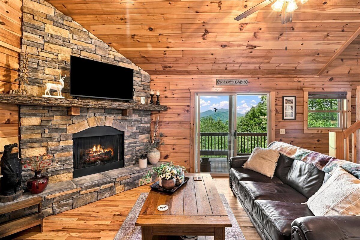 A cozy cabin interior with a stone fireplace, leather sofa, and a large window offering a view of the outdoors.