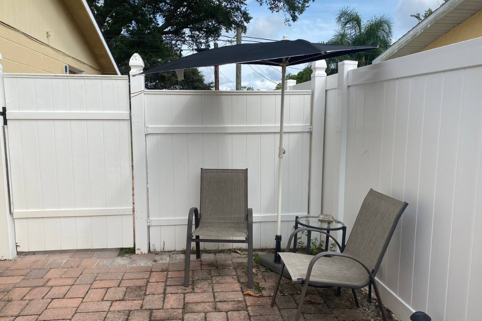 A patio area with a white fence, two chairs, a small table, and a black umbrella.
