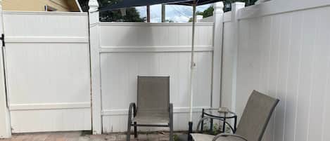 A patio area with a white fence, two chairs, a small table, and a black umbrella.