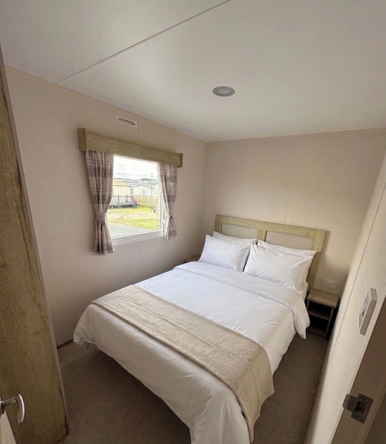 A single-bed room with a wooden headboard, a window with curtains, and a door on the left.