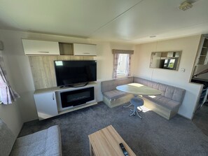A compact living space with a flat-screen TV, a sofa, a small table, and a remote control on a wooden surface. - Explore Flamborough & Beyond: Comfy Caravan on Action-Packed Haven Site (Flamborough)