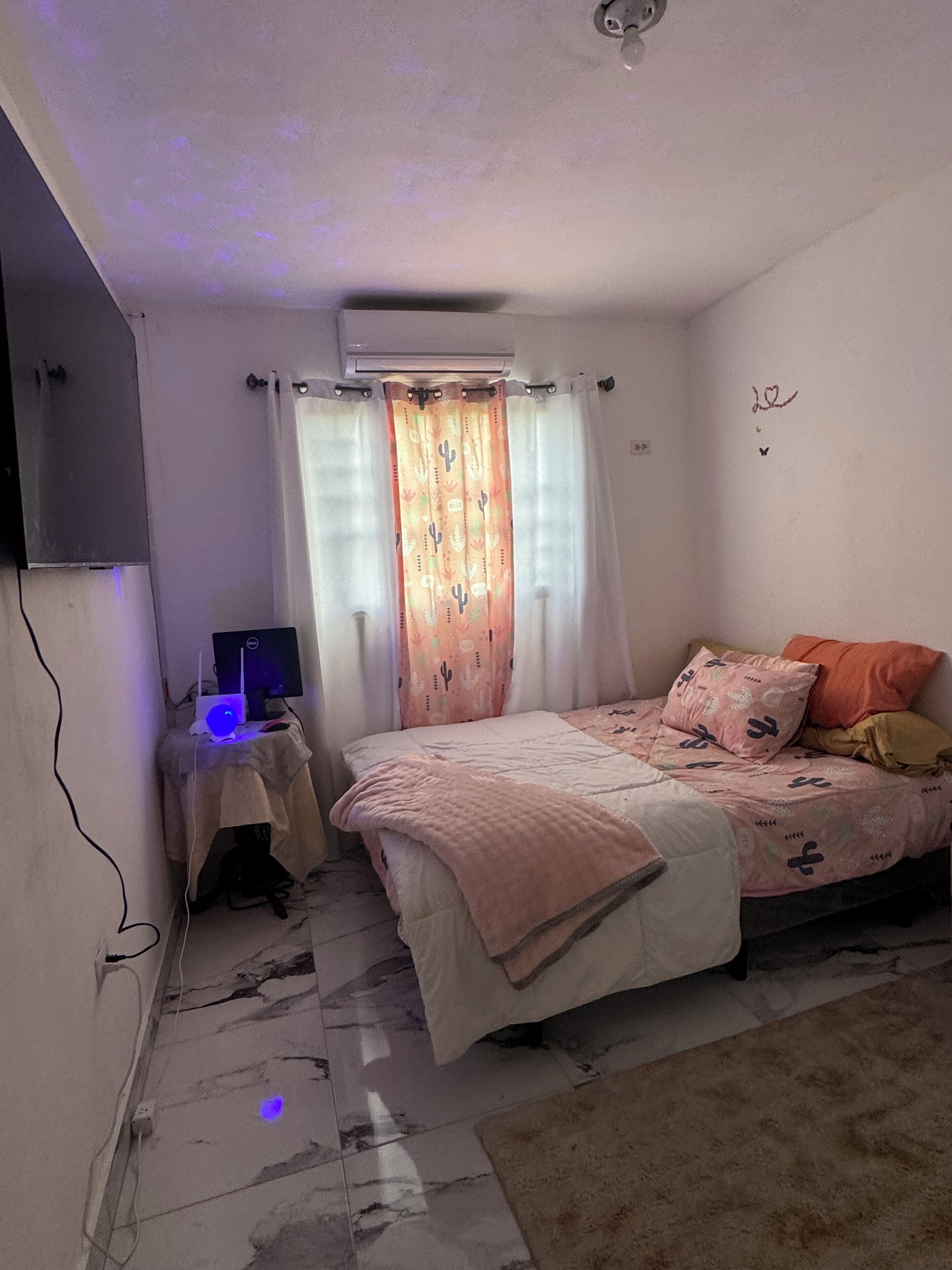 Ein Schlafzimmer mit einem Bett, einem Schreibtisch mit Computer, einer Klimaanlage und einem Fenster mit Vorhängen.