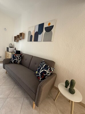 A modern living room with a grey sofa, abstract wall art, a small white table with cacti, and a kitchen area in the background. - Sisters Sardegna. Studio flat. Near Cagliari and on the coast. Pet Friendly. (Quartu Sant'Elena)