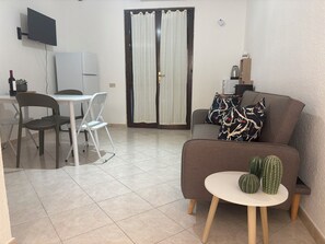 A living room with a sofa, a small round table with decorative items, a dining area with chairs and a table, a television mounted on the wall, and a sliding door with curtains. - Sisters Sardegna. Studio flat. Near Cagliari and on the coast. Pet Friendly. (Quartu Sant'Elena)