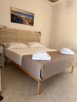 A neatly made bed with a brown quilt, two pillows, and folded towels on a wooden bed frame in a room with a simple design. - Sisters Sardegna. Studio flat. Near Cagliari and on the coast. Pet Friendly. (Quartu Sant'Elena)