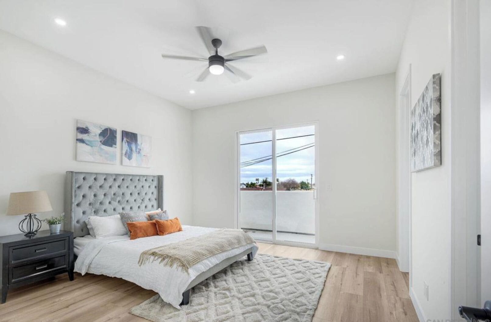 A bedroom with a tufted headboard, a bed with white and orange bedding, a bedside table with a lamp, a ceiling fan, and a view through a sliding glass door.