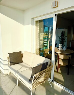 Ein Balkon mit einer weißen Couch, einem Korbstuhl und einer Glastür, die in einen Innenbereich mit Tisch und Stühlen führt.