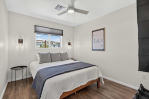 A bedroom with a bed, a ceiling fan, a window with blinds, a wall-mounted light, and a framed picture. - Iron Horse at Hoff & Co | Off 4th Ave (Tucson)