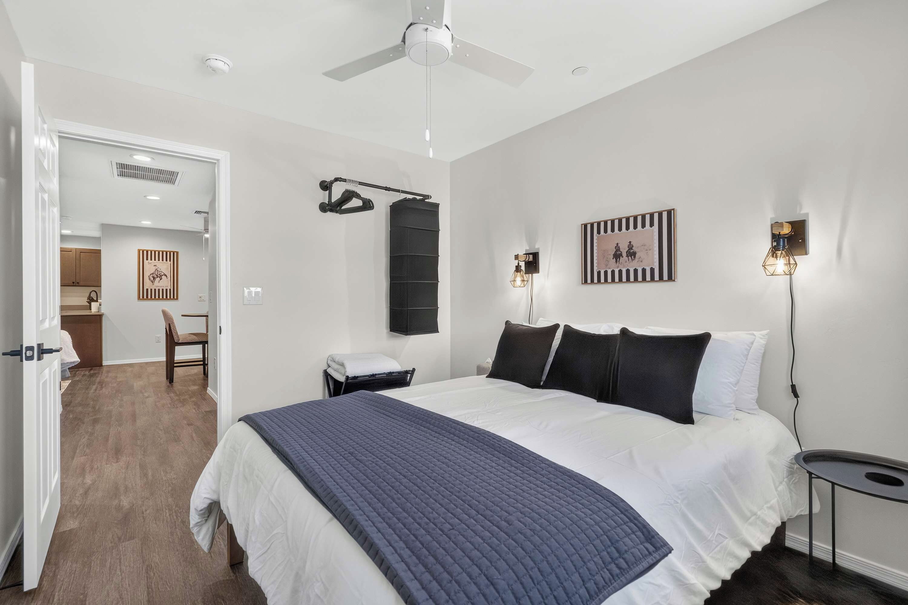 Une chambre à coucher comprenant un lit, des tables de chevet, un ventilateur de plafond, une lampe fixée au mur et une porte donnant accès à une salle de bain.