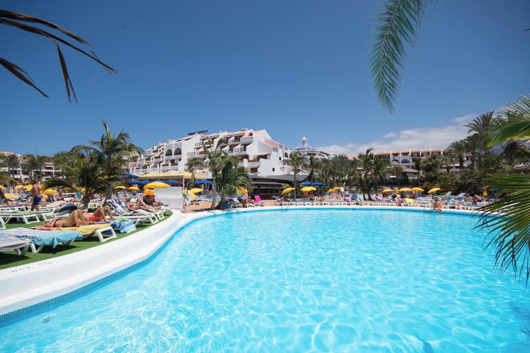 A swimming pool with lounge chairs and umbrellas, surrounded by palm trees and a hotel building with multiple balconies.