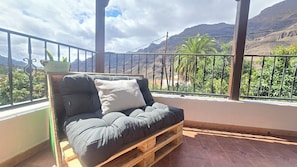 Ein Balkon mit einer Holzpaletten-Couch, einem kleinen Tisch mit einer Pflanze und Blick auf Berge und Grün.