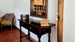 Ein Zimmer mit Holzboden, einem dunklen Holztisch mit Büchern, einem Korbstuhl, einem Spiegel und einem Kleiderständer.