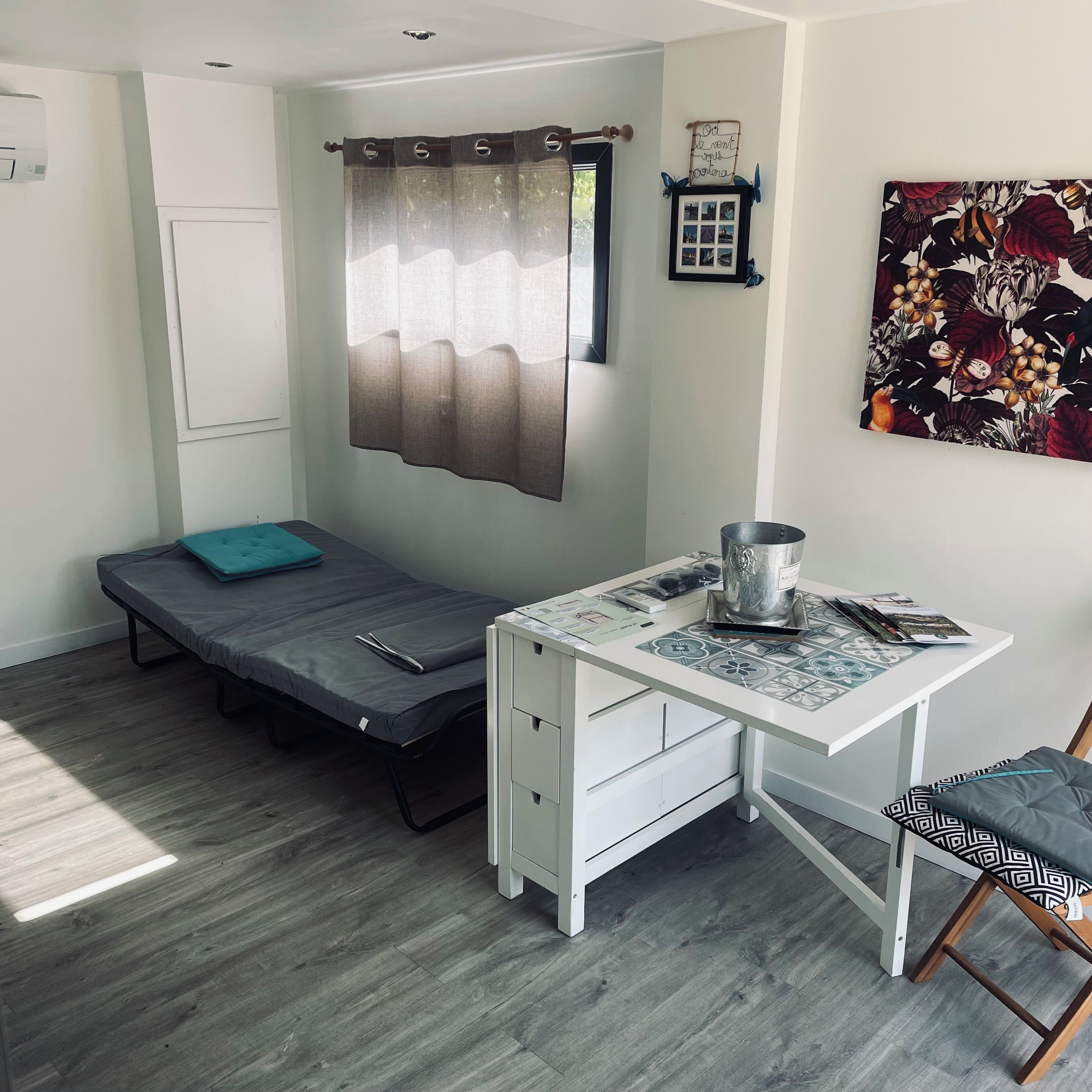 Een knus ingerichte kamer met een bed, een bureau met stoel, een klein tafeltje met waterkoker, en een wandplank met decoratieve objecten.