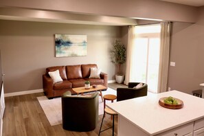 A modern living room with a brown leather sofa, a coffee table, and a dining area with a white countertop and a bowl of green apples.