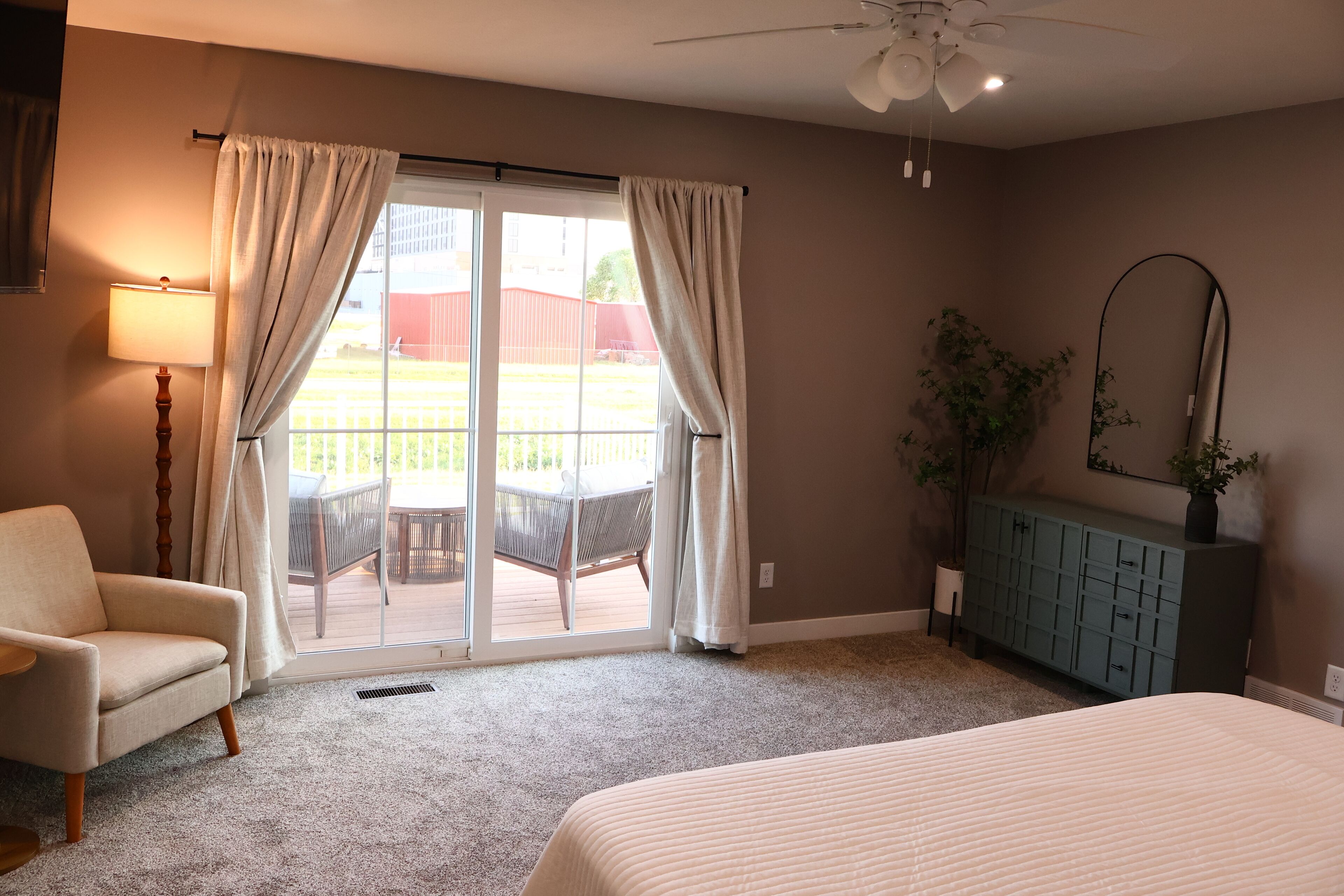 A bedroom with a bed, a chair, a desk, a lamp, a ceiling fan, and a view of the outdoors through a sliding door.