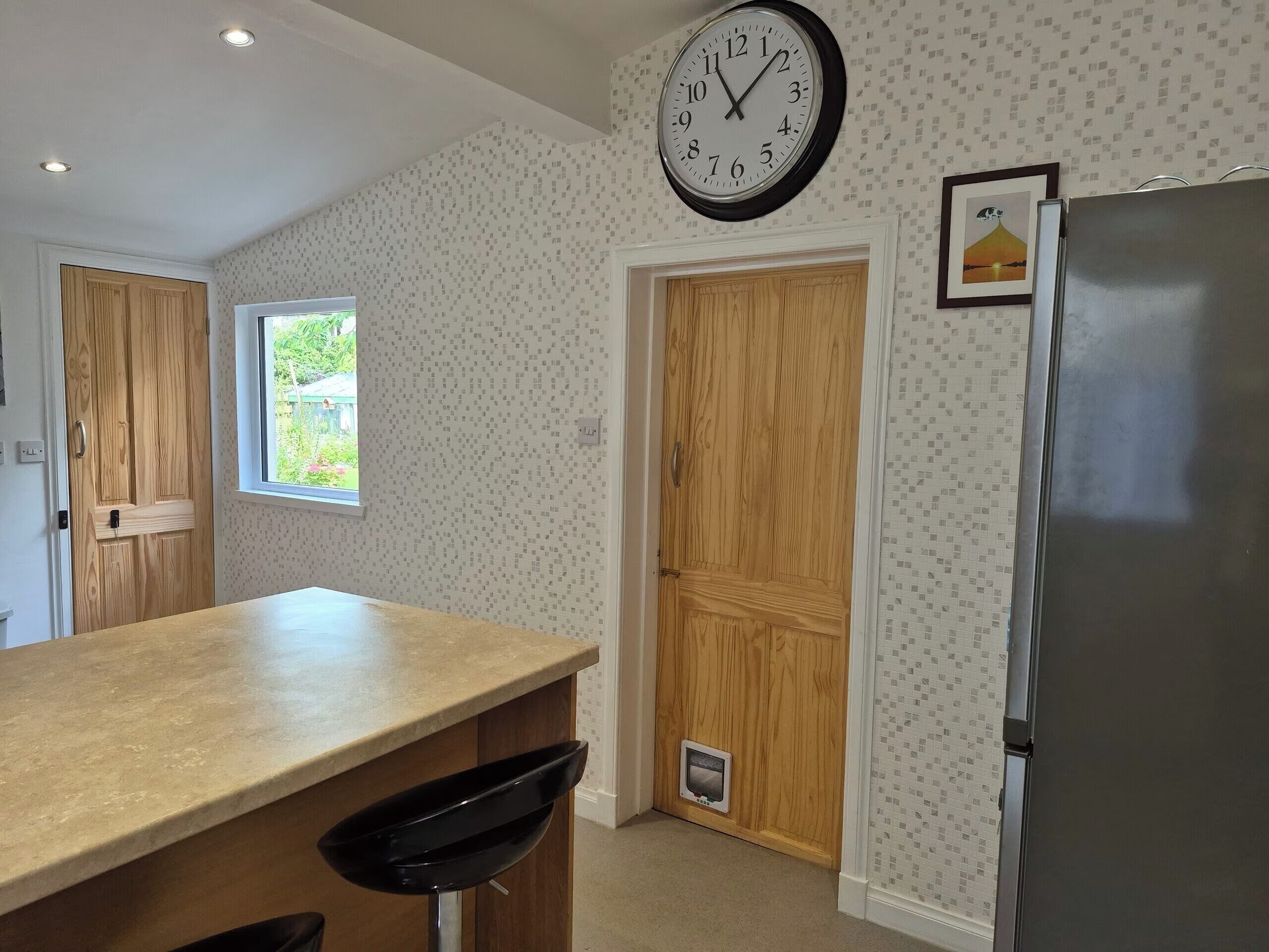 A kitchen with a wooden countertop, a black chair, a wall clock, a refrigerator, and a window with a view of greenery.