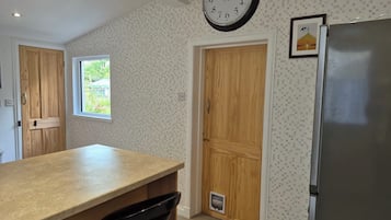 A kitchen with a wooden countertop, a black chair, a wall clock, a refrigerator, and a window with a view of greenery.