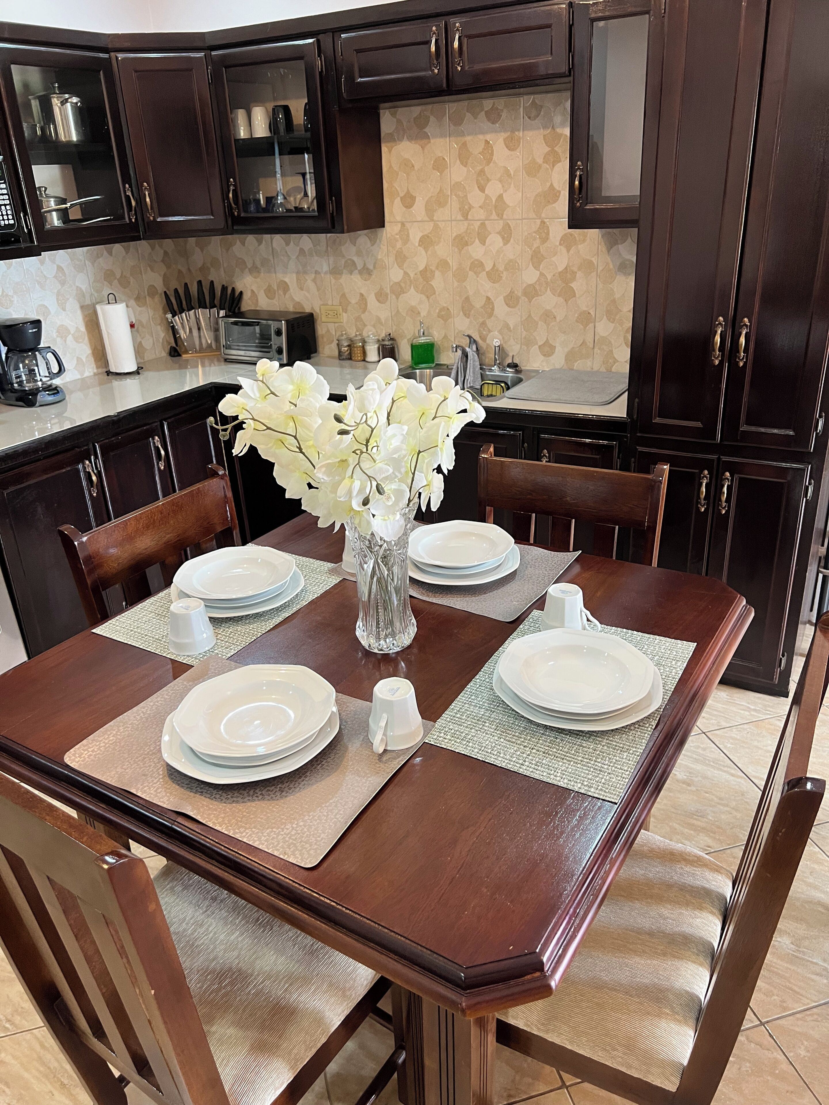 A dining table set with plates, a vase of flowers, and a coffee maker in the background.