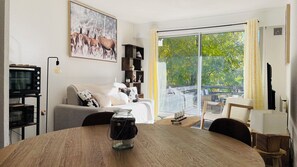 Ein gemütliches Wohnzimmer mit einer Couch, einem Couchtisch und einem Fernseher. Ein großes Fenster bietet Blick auf Bäume.