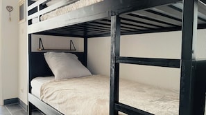 A bunk bed with a white mattress and a pillow, a shelf above the lower bunk, and a wall-mounted light fixture. - Cozy Studio (Formiguères)