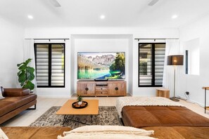 Ein modernes Wohnzimmer mit Flachbildfernseher, Holzmöbeln und einem großen Fenster mit Jalousien.