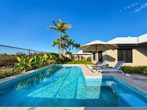 A rectangular swimming pool with clear blue water, surrounded by a fence, palm trees, and a building with a sloped roof.